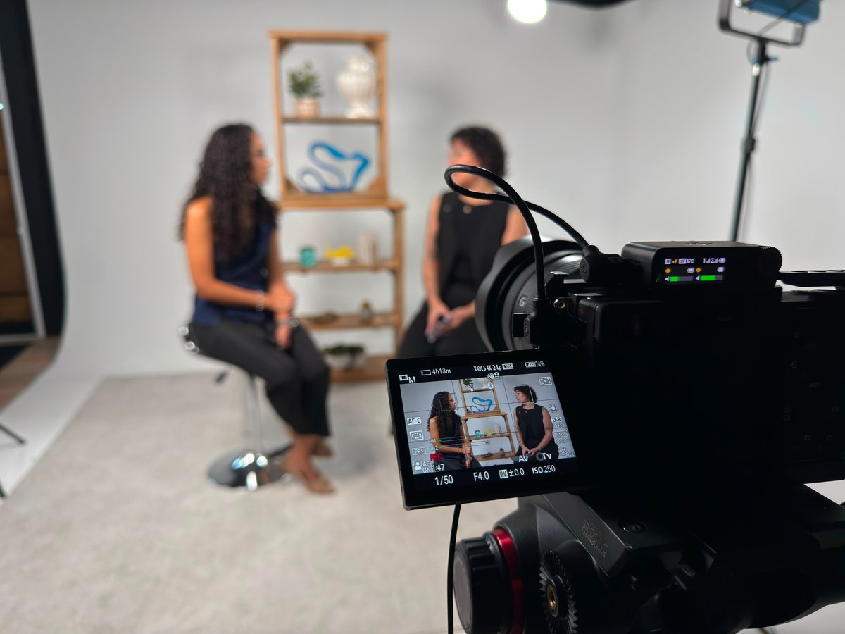 Video camera recording two people in a studio setting with shelving and professional lighting equipment visible in background