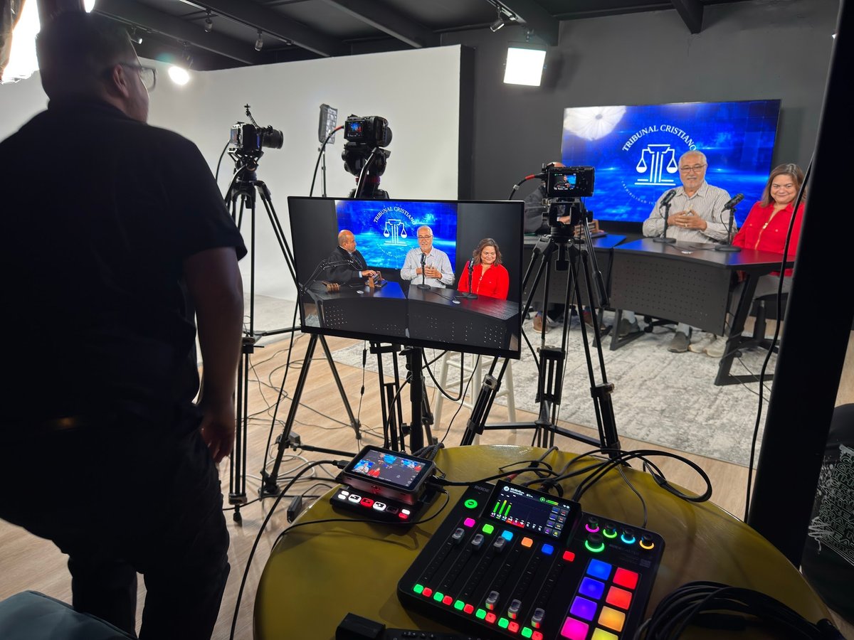 Television studio production setup with camera operators, mixing console in foreground, and live broadcast of news or talk show on monitors
