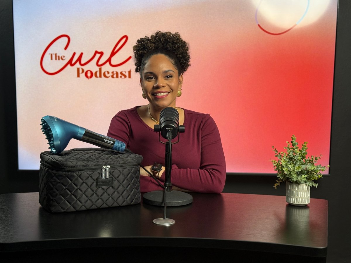 Woman smiling at microphone in front of The Curl Podcast backdrop with quilted bag and small potted plant on desk