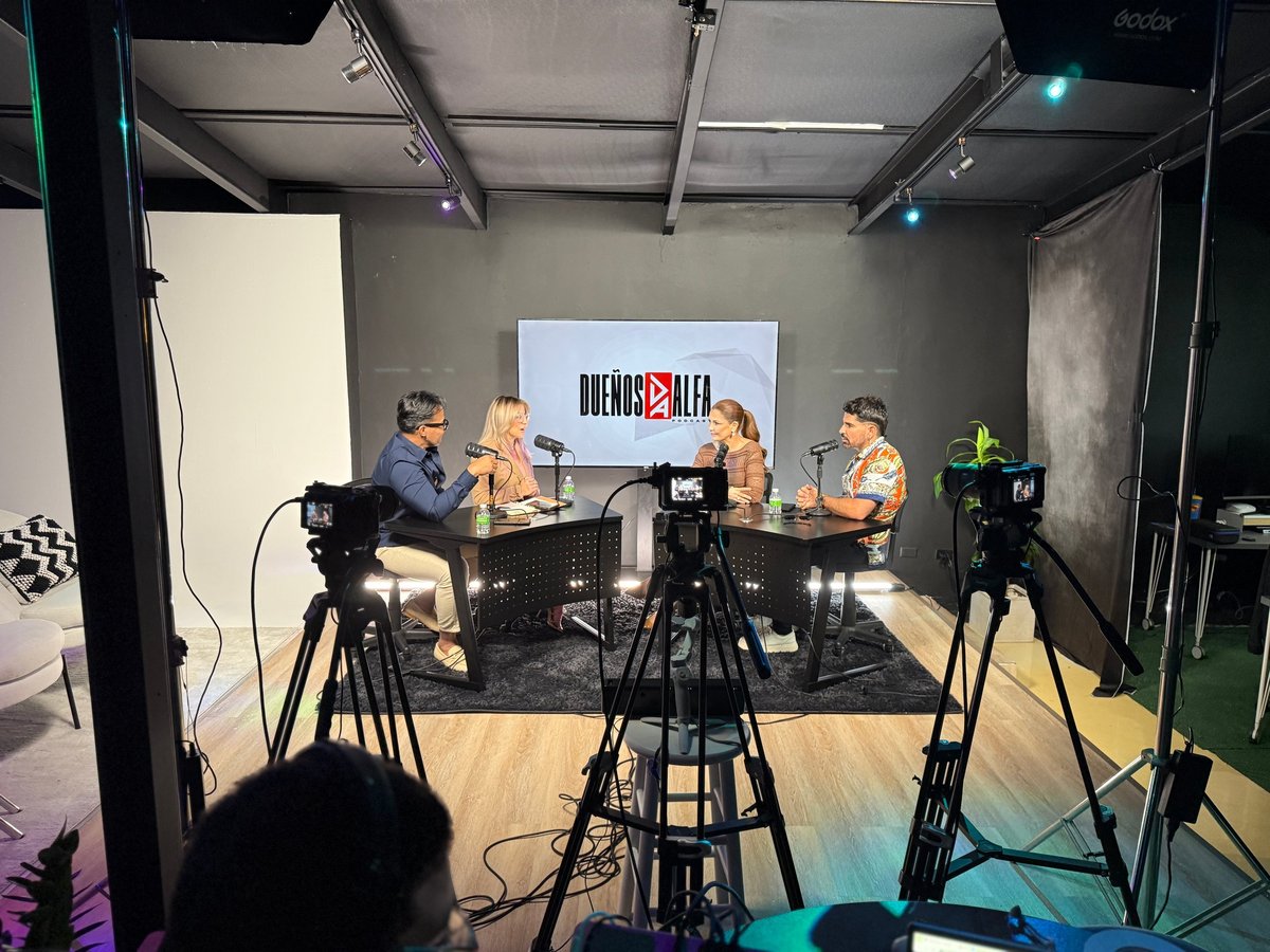 Podcast or talk show studio recording with four people seated at table, multiple cameras filming, professional lighting setup