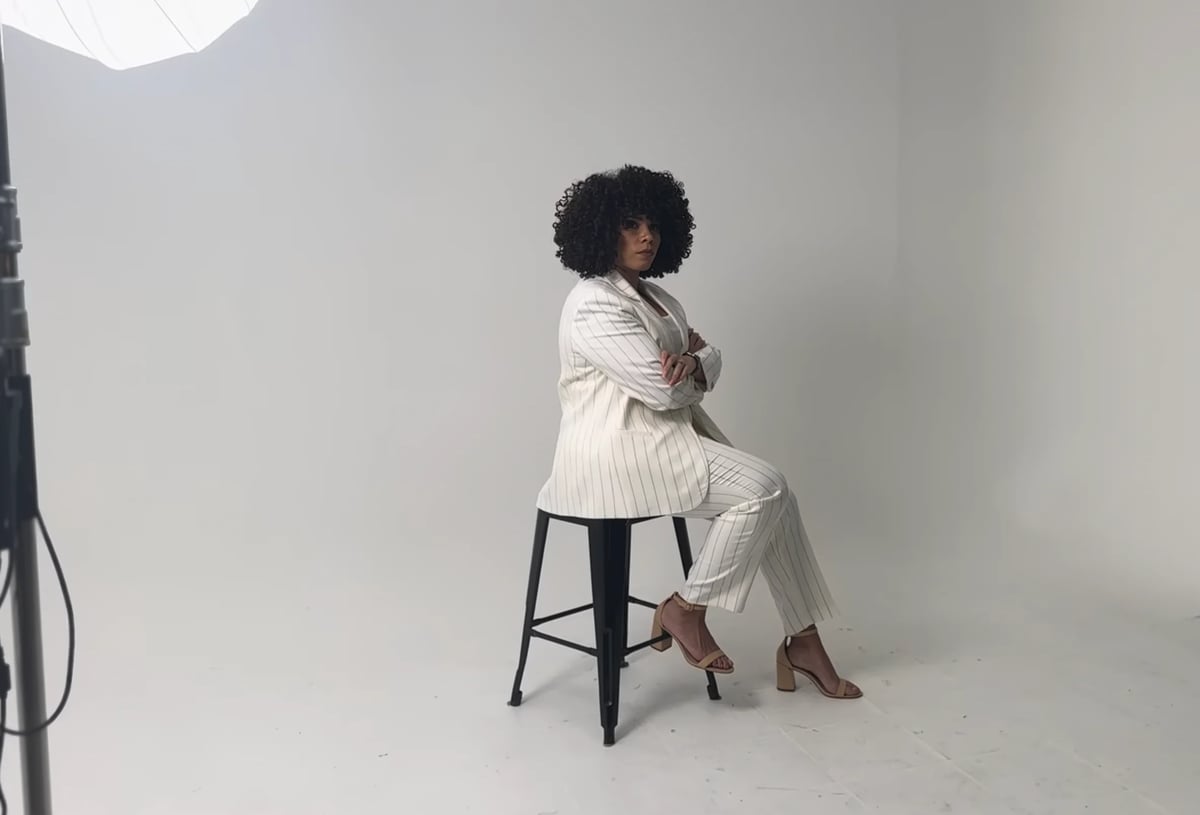 Woman with curly black hair sitting on a black stool against a white background, wearing a cream-colored quilted jacket